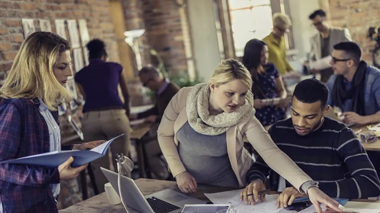 Three people working at a desk with other people in the background working at other desks